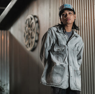  Model wearing ATH Good Art Mack grey washed Chore Coat and grey and black logo trucker hat at the Good Art Foundry space