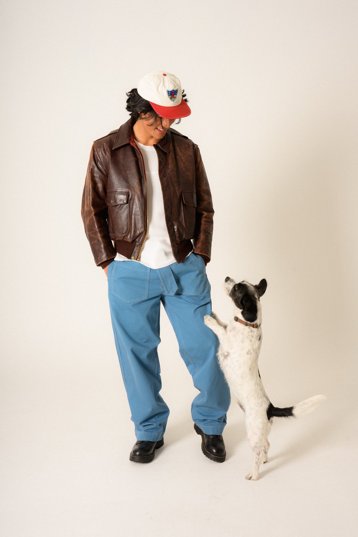 Man wearing warm indigo blue belted moto pants in brown leather jacket with hands in pockets looking down at a dog