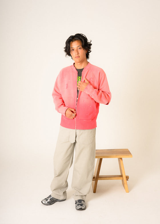 Person wearing a pink jacket and beige pants standing next to a wooden stool on a white background