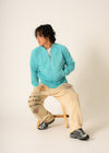 Person wearing a turquoise jacket and beige pants sitting on a chair against a white background