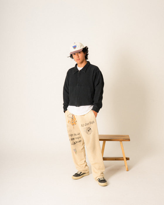 Person wearing a black jacket, natural color pants, and a two tone cap standing next to a wooden bench on a plain background