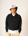 Person wearing a black jacket, white shirt, and beige pants with a cap on a plain background