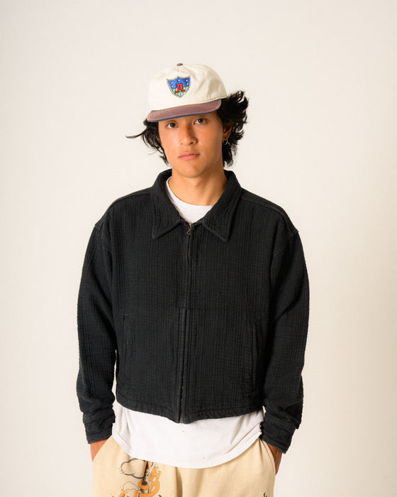 Person wearing a black jacket, white shirt, and beige pants with a cap on a plain background