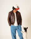 Person wearing a brown leather jacket and blue pants standing next to a small white and black dog on a plain background