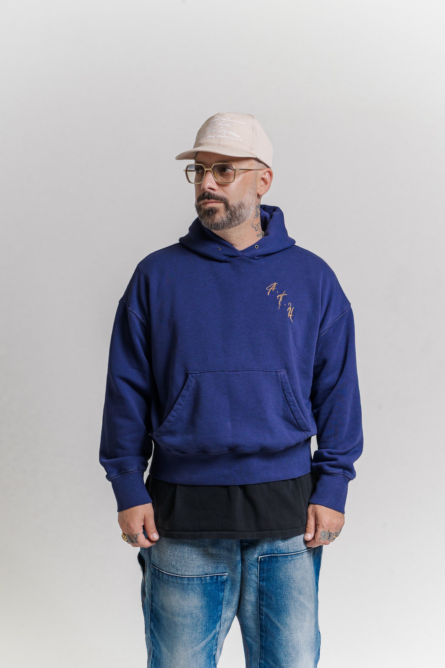 Man wearing a blue hoodie with text, glasses, and a cap on a plain background
