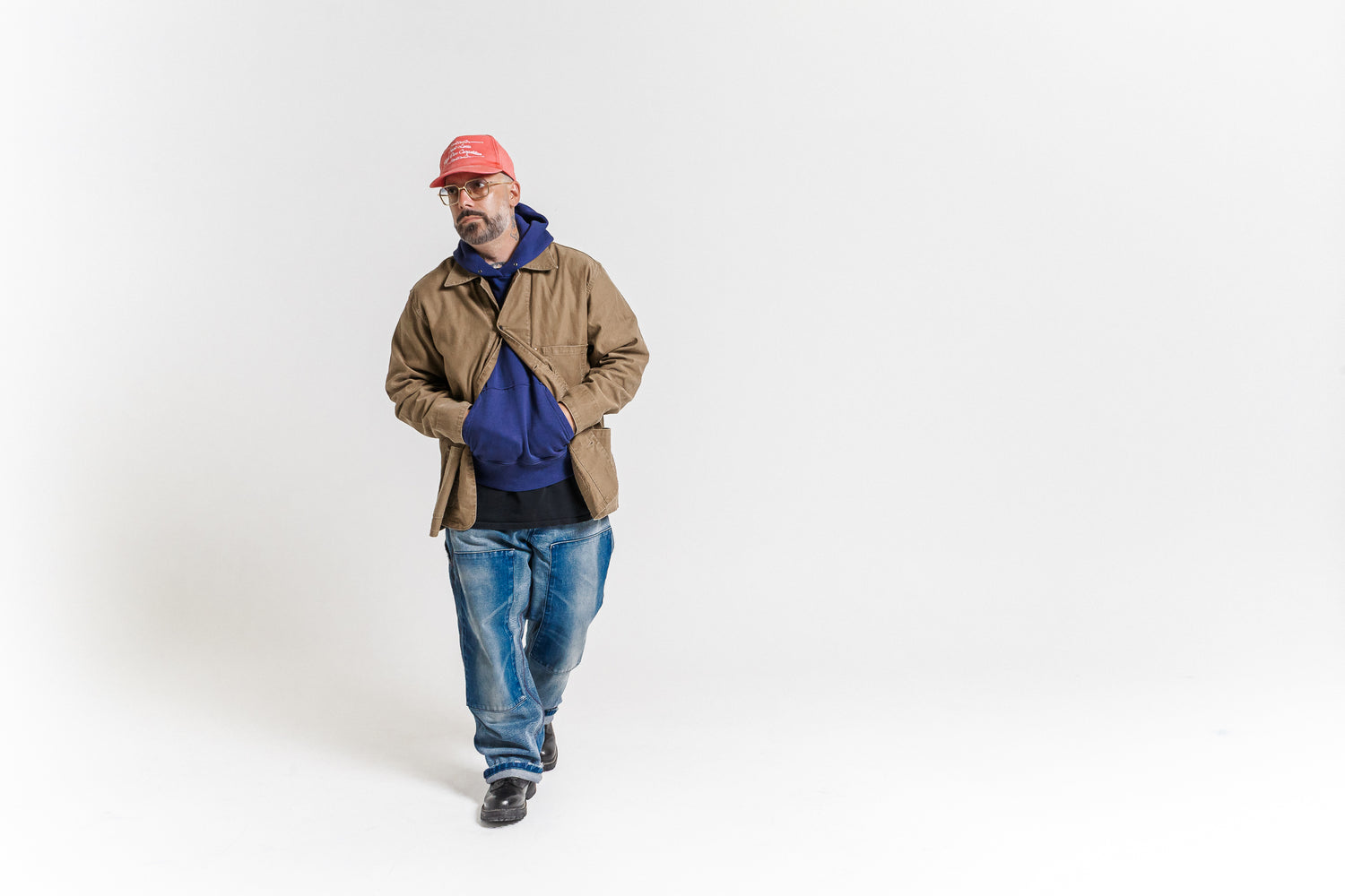 Man wearing a brown jacket, blue jeans, and a red cap on a white background