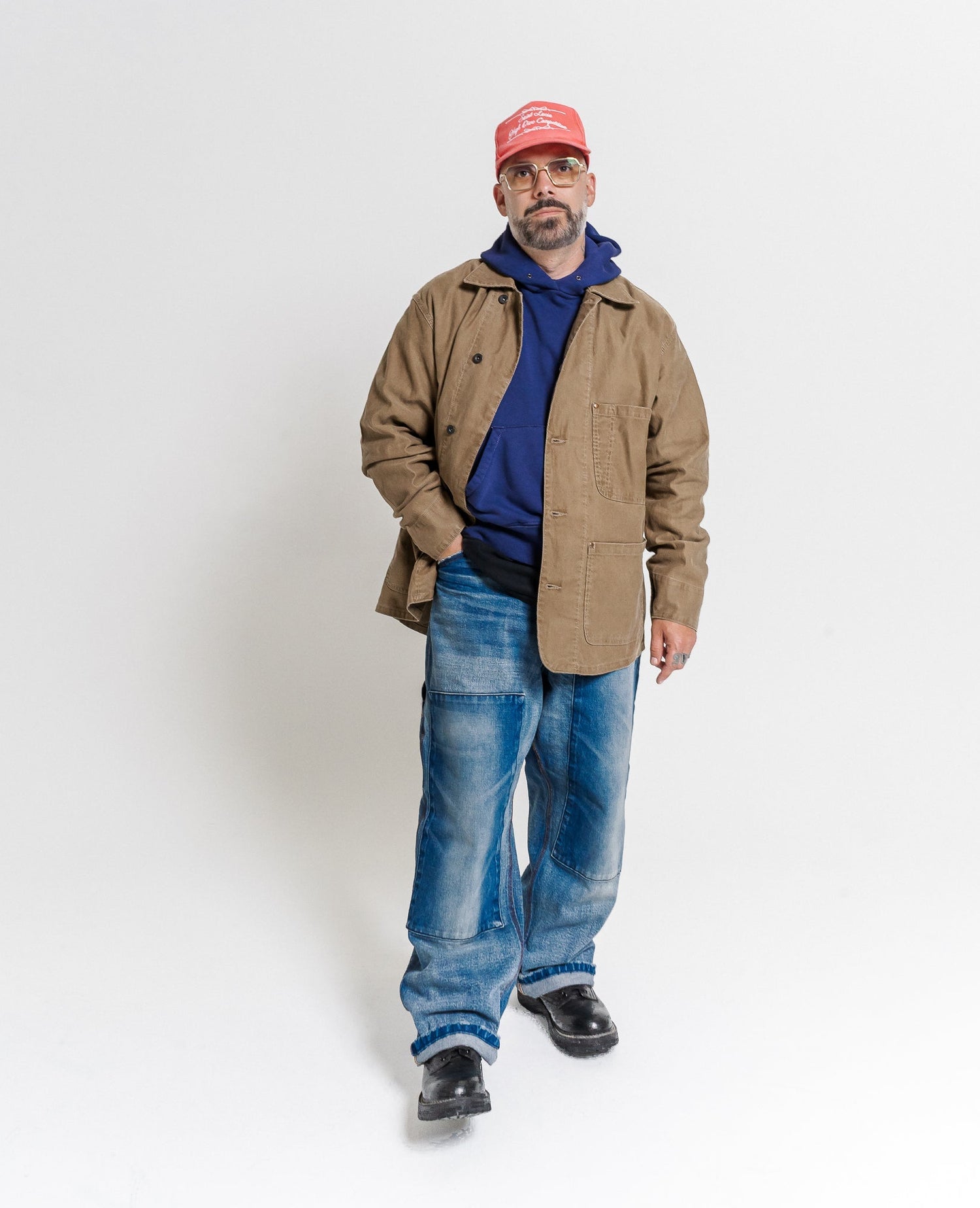 Man wearing a tan jacket, blue jeans, and a red cap on a white background